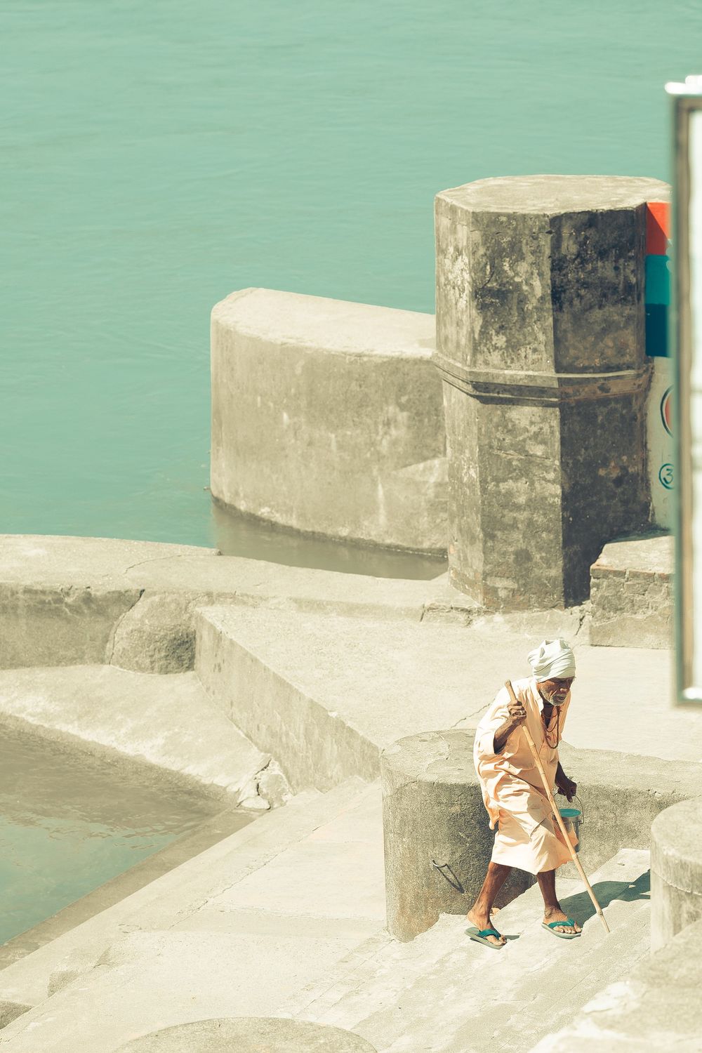 An elderly person walks with a stick along concrete steps beside turquoise water.