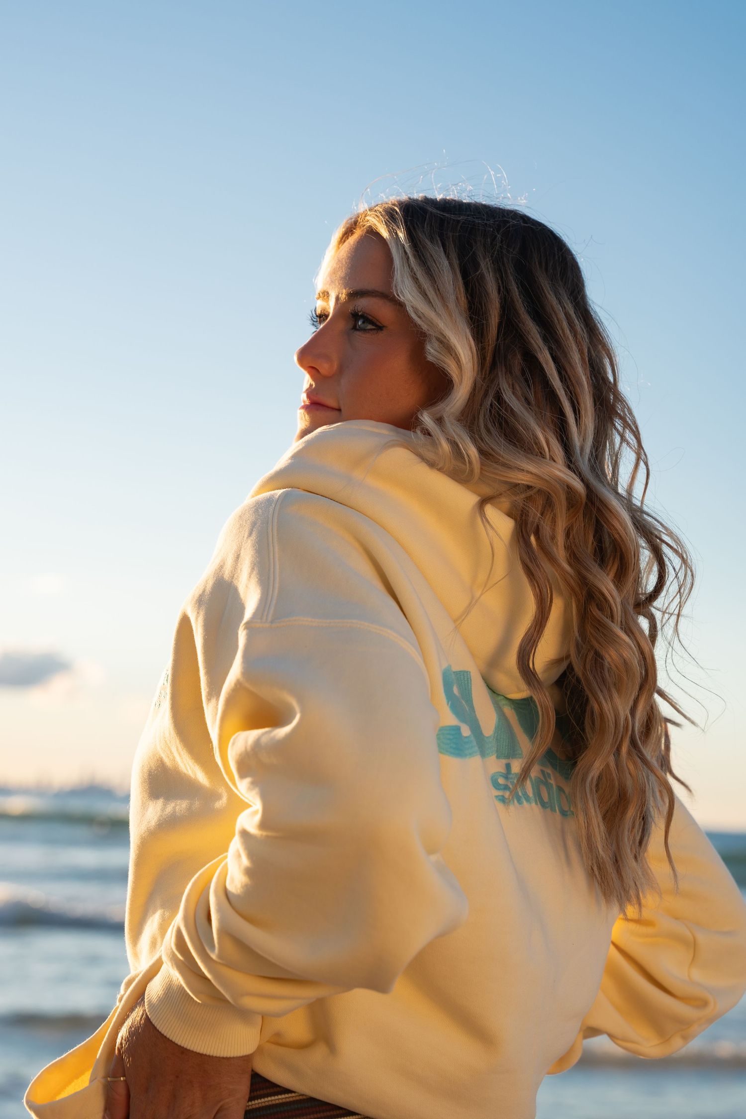 Woman in cream hoodie with teal logo poses at beach during golden hour with wavy blonde hair.