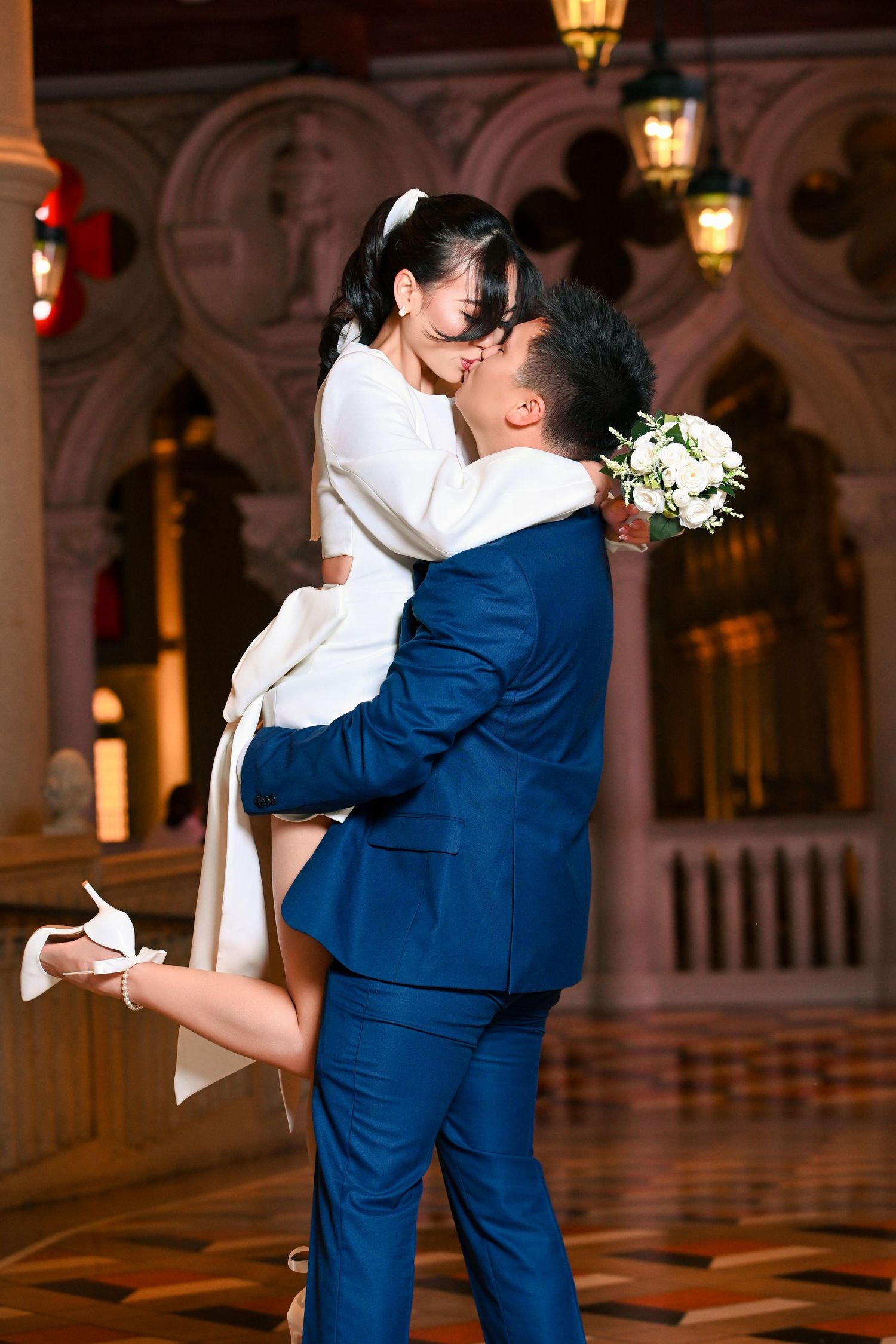 Groom in blue suit lifts bride in white dress for a kiss, holding bouquet, in elegant venue.
