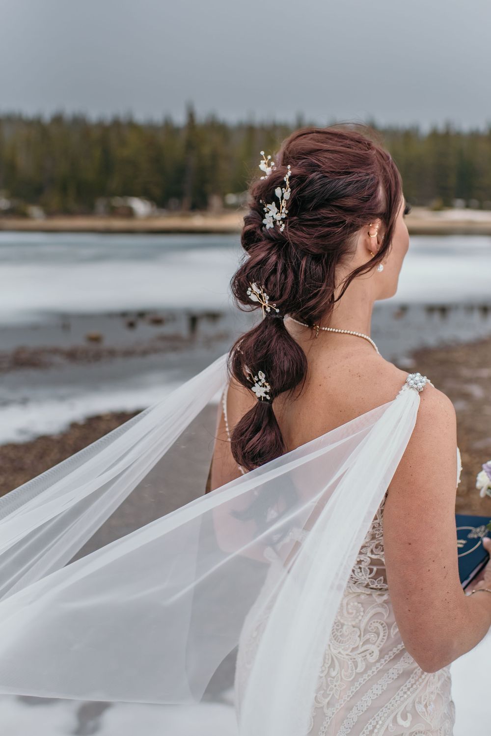 Bride with auburn braided updo adorned with floral pins, white lace gown and flowing veil outdoors.