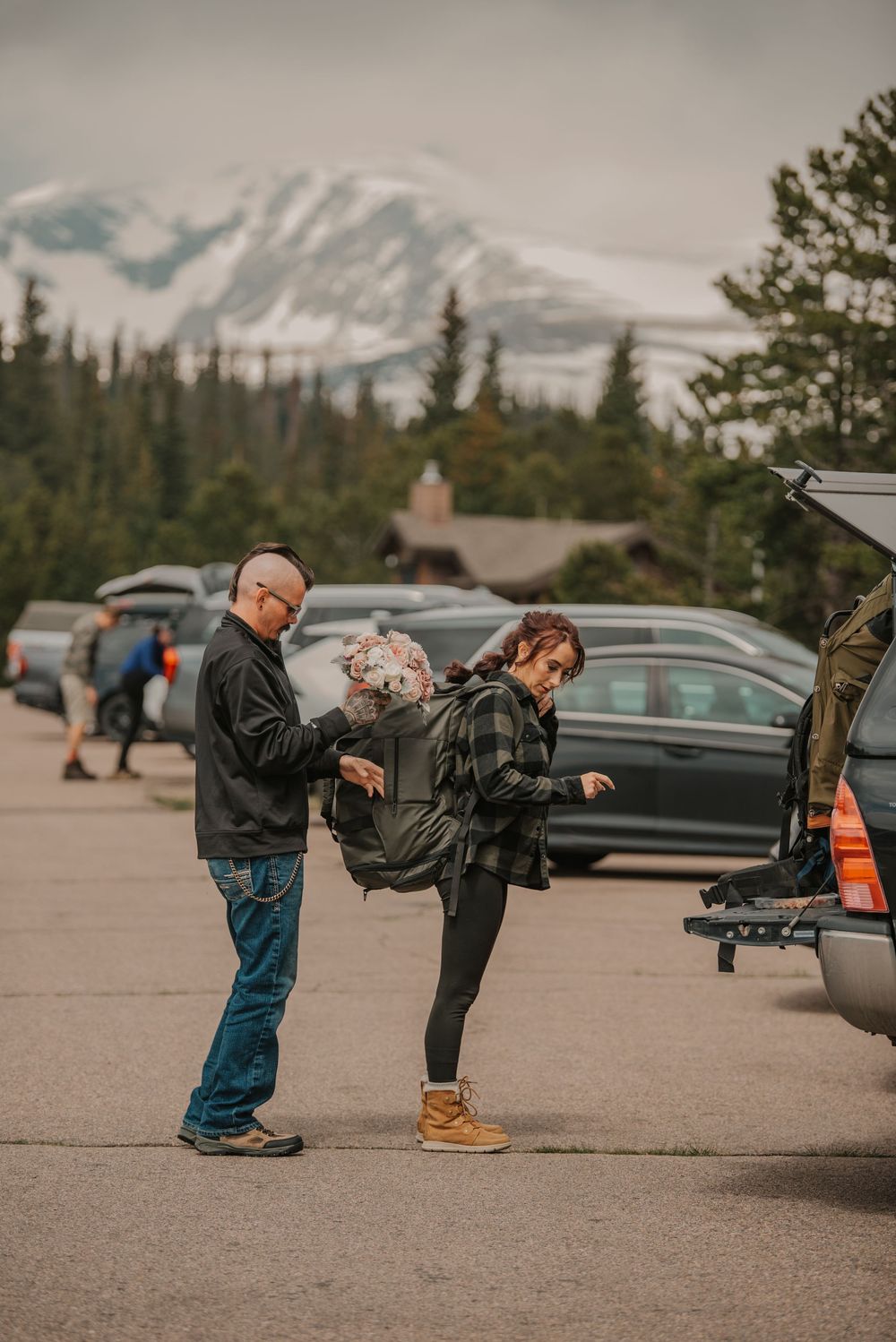Couple prepares hiking backpacks in mountain parking lot with snow-capped peak visible in background.