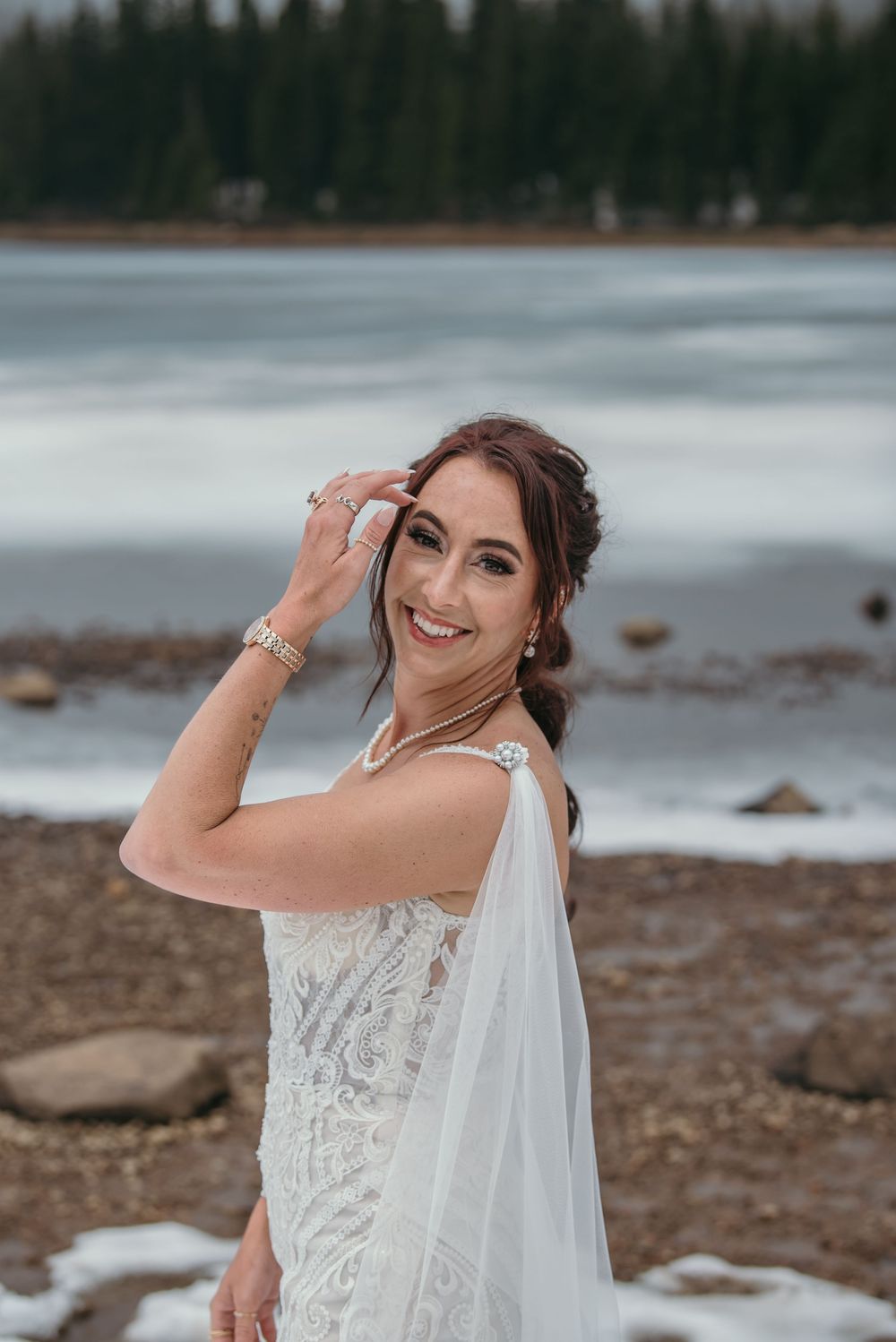 Smiling bride in lace wedding gown and veil posing by a frozen mountain lake on her wedding day.