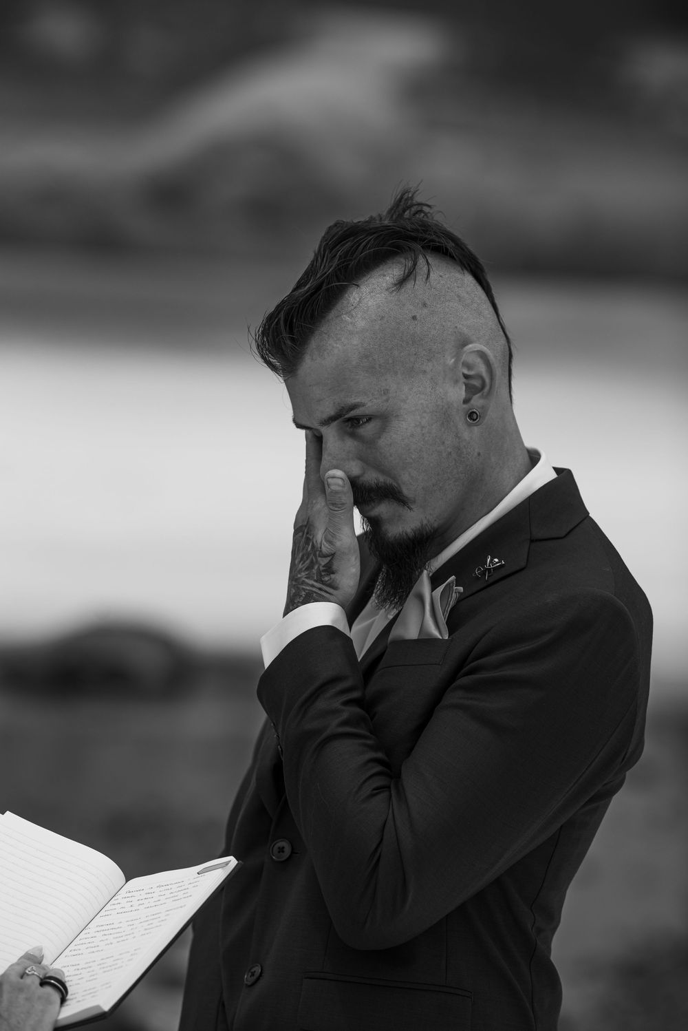 Emotional groom in black suit wiping tears while reading wedding vows from a book in black and white photo.