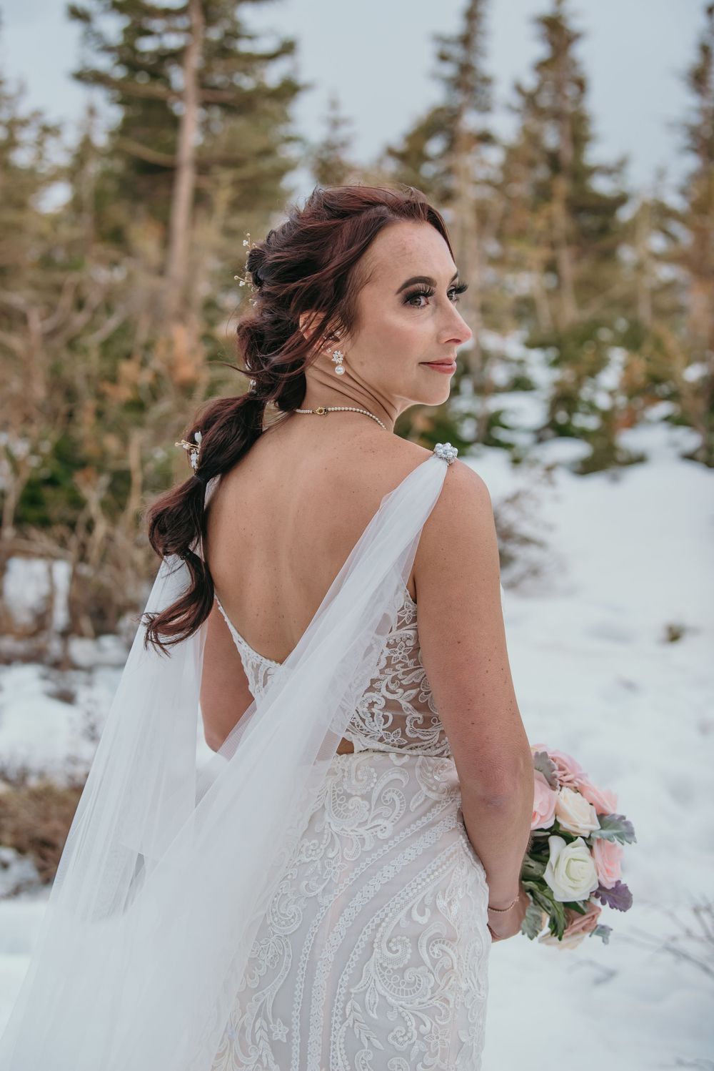 Bride in backless lace wedding gown holding bouquet in snowy forest setting.
