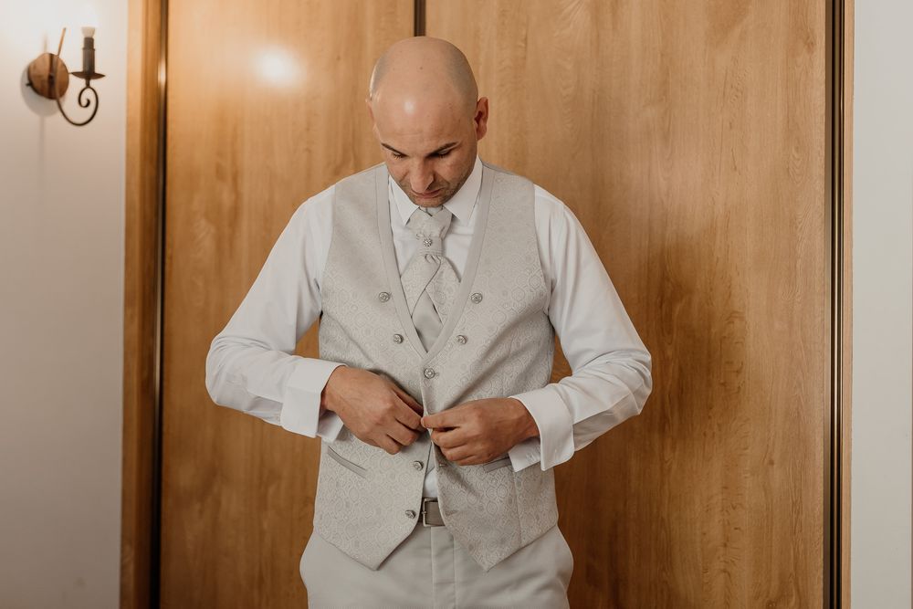 Bald groom buttoning his light gray vest while getting ready for his wedding ceremony.