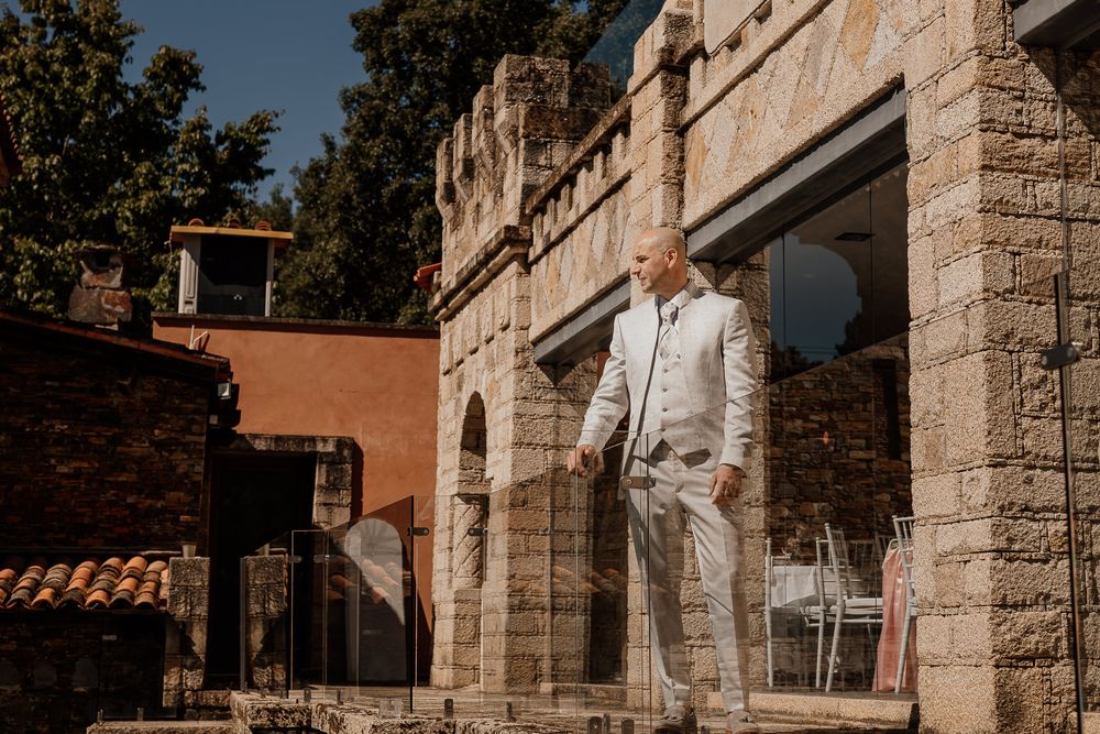 Man in light gray suit standing on stone steps of a rustic Mediterranean-style building on a sunny day.