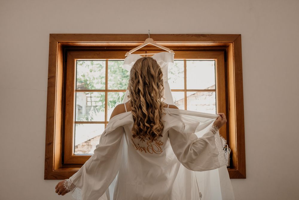 Bride in white robe holding wedding dress on hanger in front of rustic wooden window.
