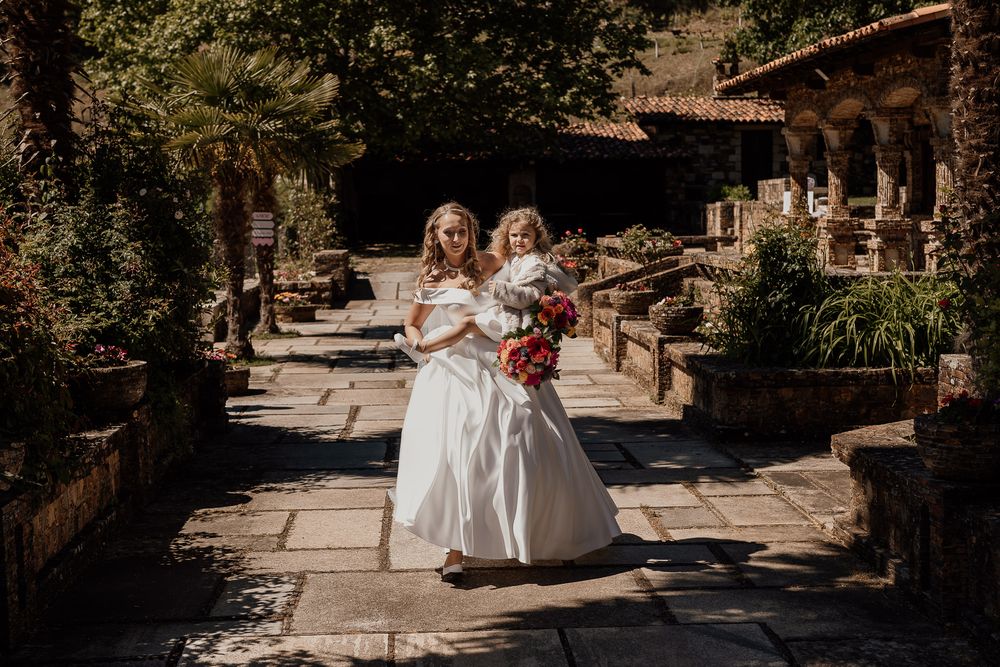 Two brides in white gowns walk together on a stone path, one holding a vibrant bouquet, at a rustic garden venue.