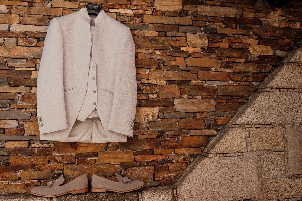 Beige linen suit jacket with vest hanging on brick wall beside tan loafers, elegant groom wedding attire display.