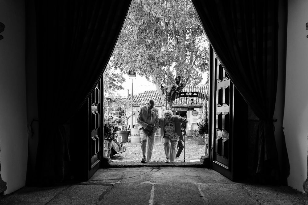 Black and white photo of two people walking through large open doors toward a sunlit courtyard with trees.