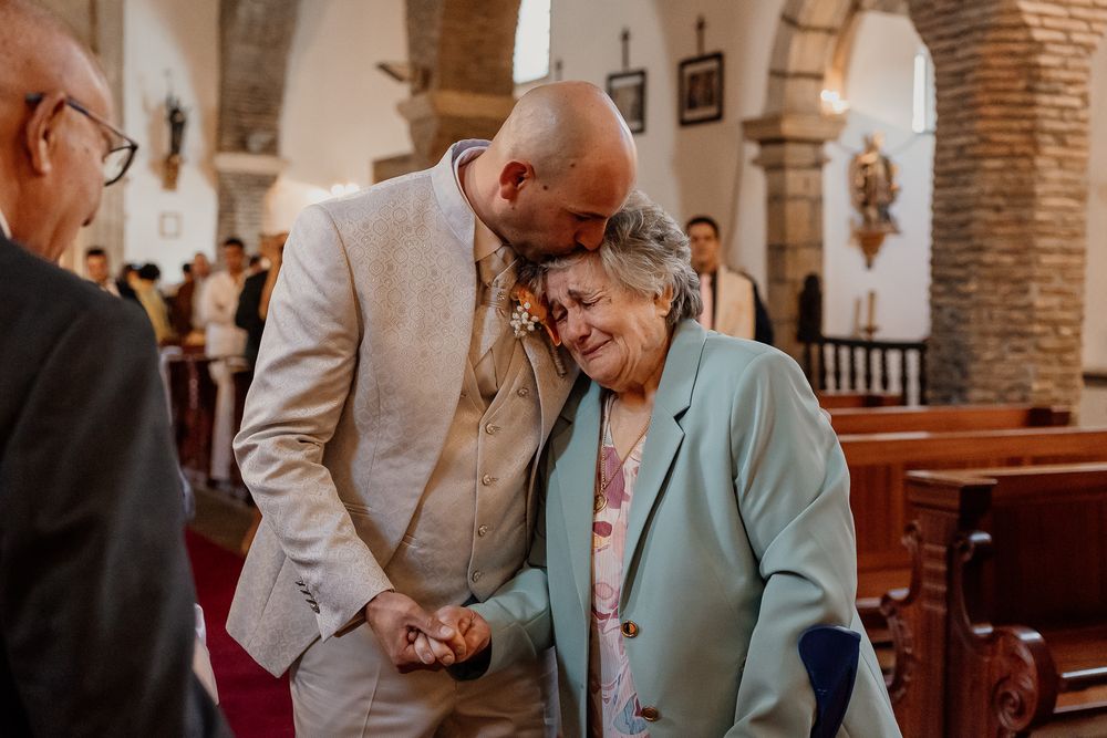 A bald groom in a beige suit shares a tender moment with an elderly woman in a church aisle.