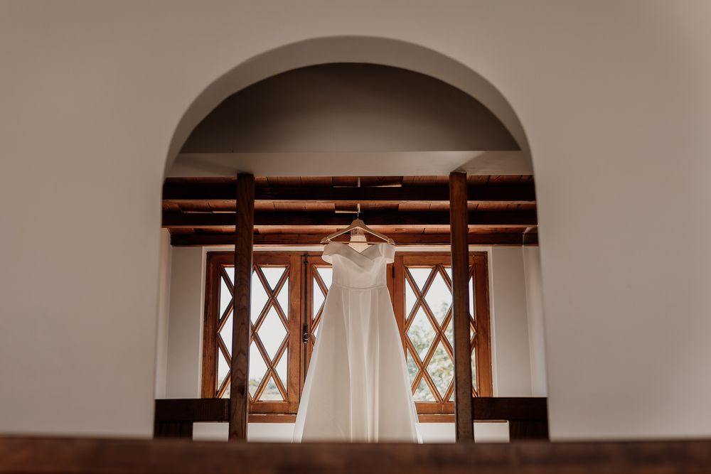 Elegant white wedding dress hanging in arched window with wooden frames, soft natural light, rustic venue.