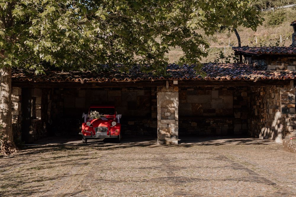 A red vintage car parked inside a rustic stone garage beneath a large tree in a rural setting.