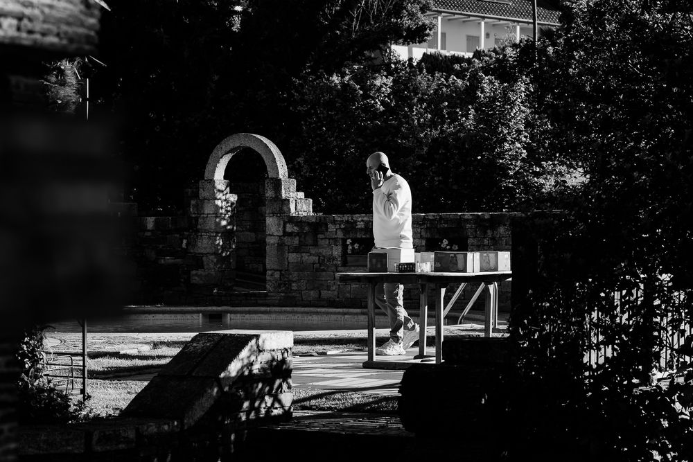 Black and white photo of a person in white coat standing at a table in a garden with stone archway backdrop.