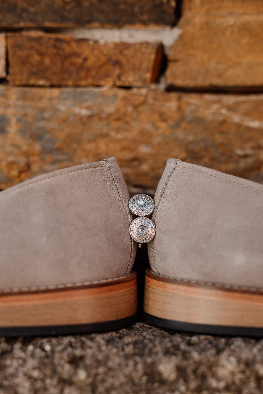 Two gray suede ankle boots with silver concho buttons displayed against a rustic brick wall backdrop.