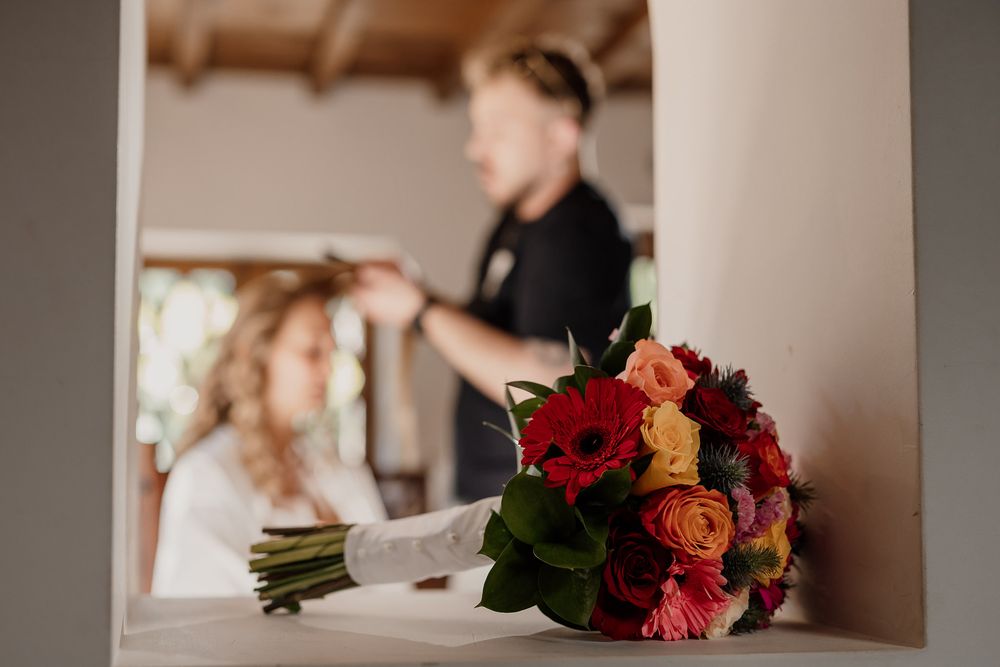 Bridal bouquet of red, orange, and pink flowers in focus, with a hairstylist working on a bride in the background.