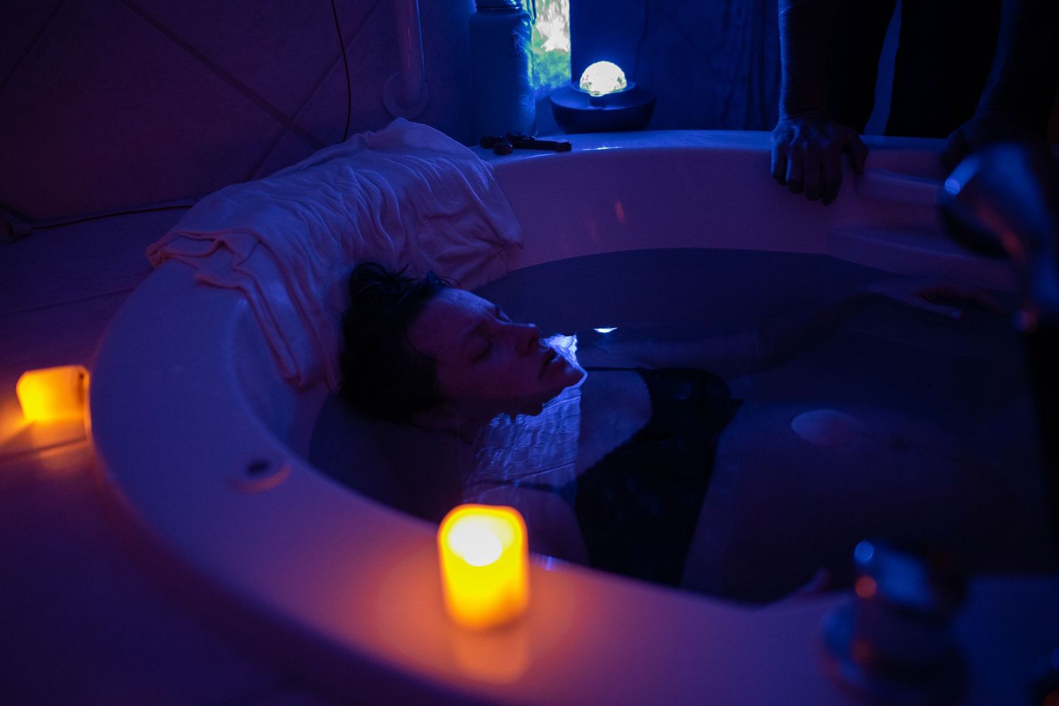Person relaxing in a round hot tub with candlelight ambiance and purple mood lighting in a luxury bathroom.