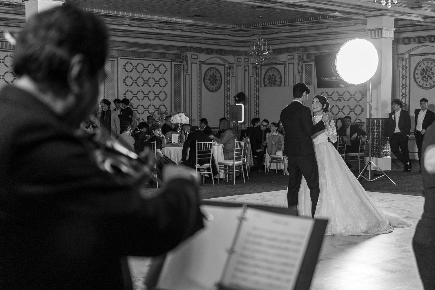 Black and white wedding photo of couple sharing first dance while violinist performs in elegant ballroom.