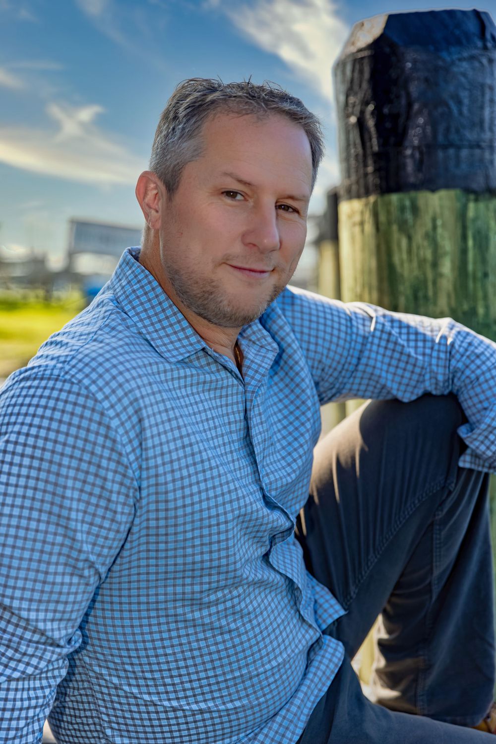 Casual waterfront portrait in blue checkered shirt with wooden posts in background.