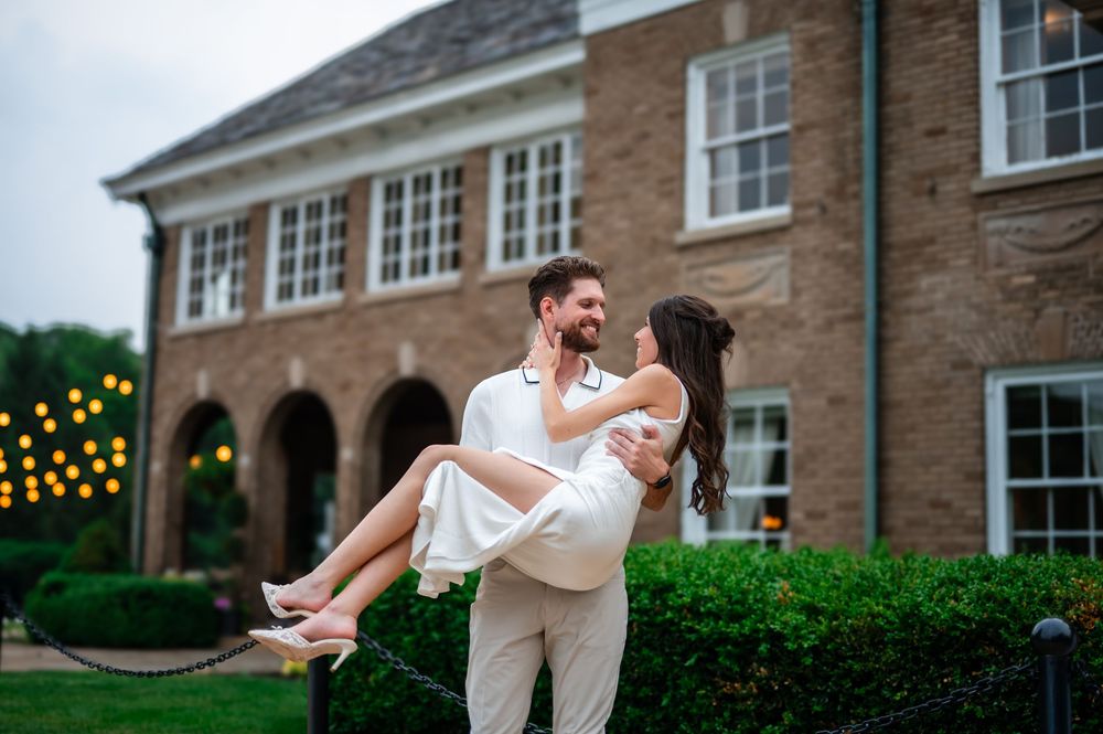 Groom lifting bride in white dress outside elegant brick mansion with string lights at dusk.