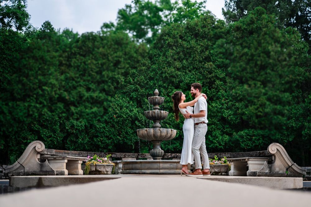 Couple embracing romantically in front of a stone fountain surrounded by lush green trees in an elegant garden setting.