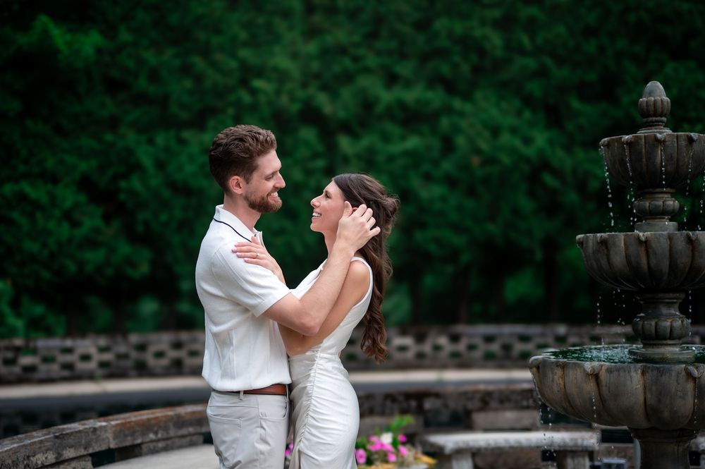 Couple in white embracing near stone fountain with lush green trees, romantic outdoor wedding portrait.
