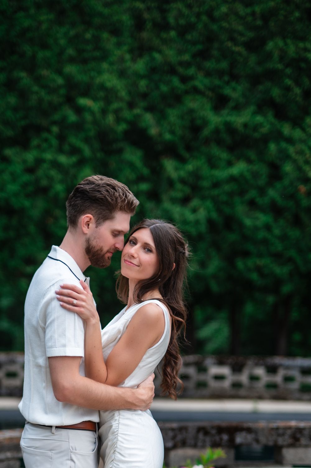 A couple in white attire embrace tenderly outdoors, with lush green foliage in the background.