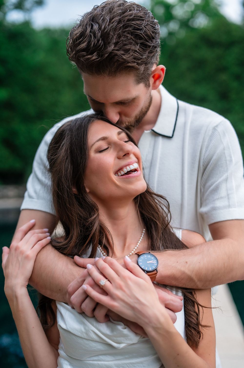 A happy couple embraces outdoors, man kissing woman's head as she laughs, both wearing white, with greenery behind.