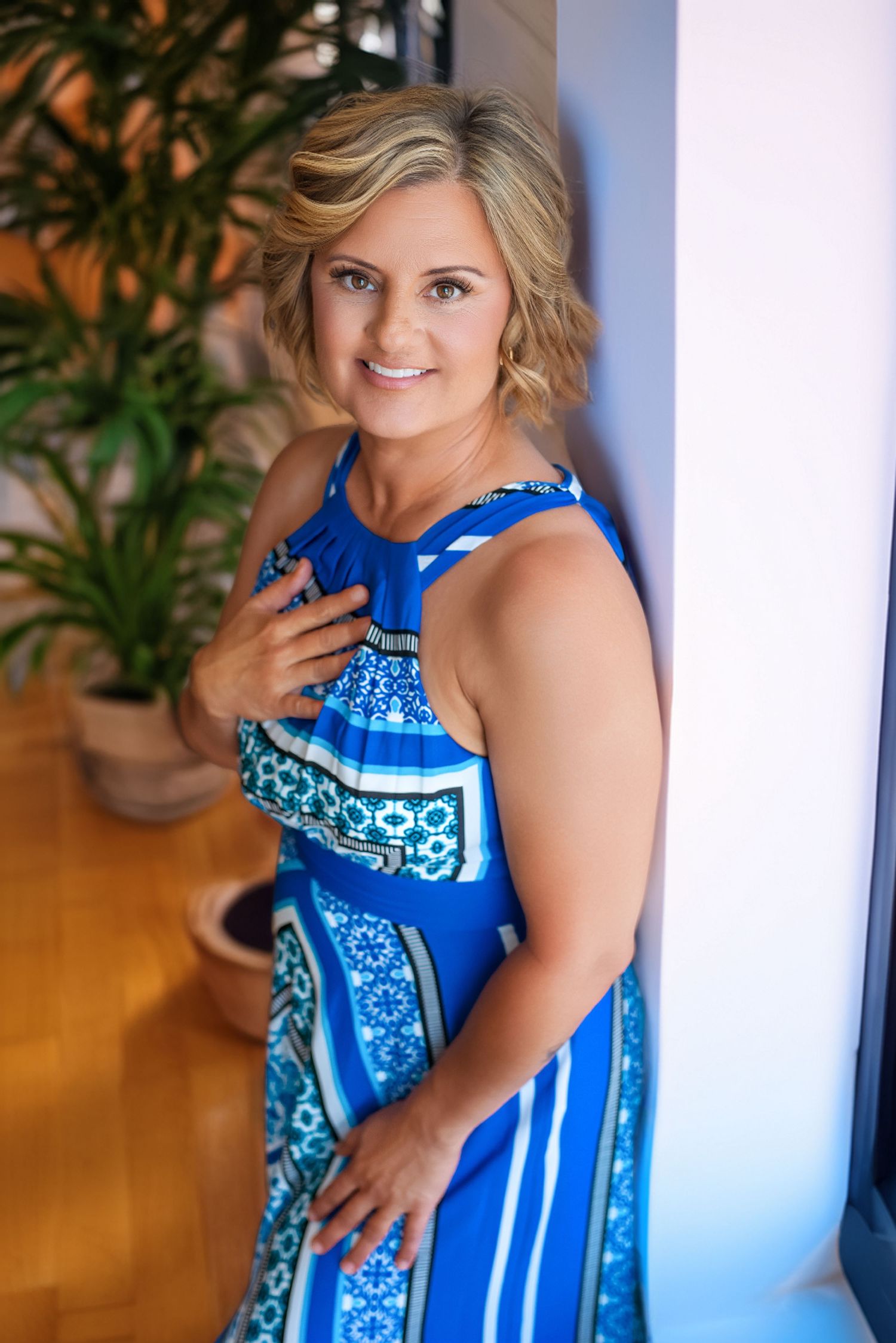 Smiling blonde woman in a blue patterned maxi dress posing gracefully against a white wall during her cape girardeau mo branding session.