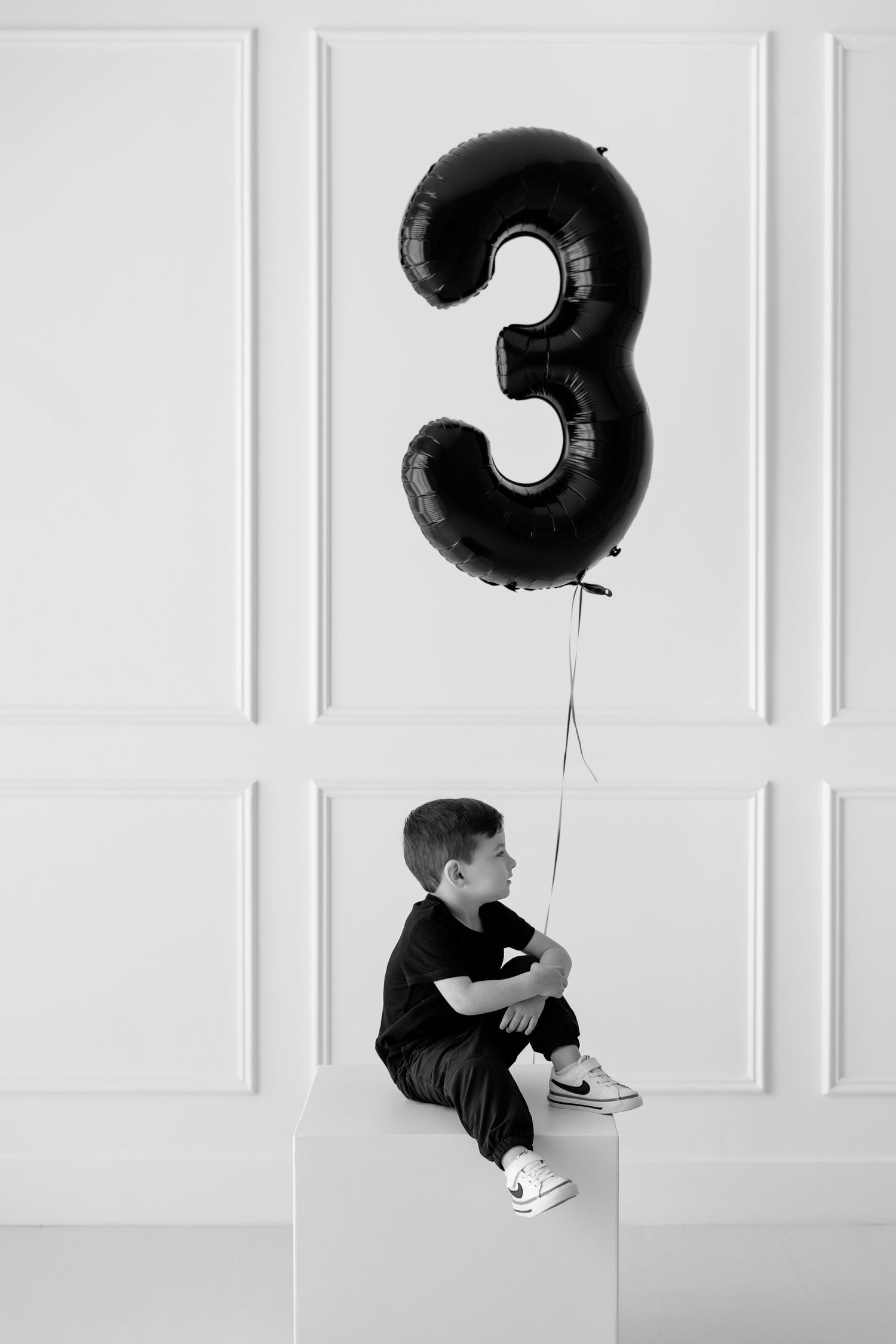Black and white photo of a young boy sitting on a white cube holding a large number 3 balloon for his 3rd birthday.
