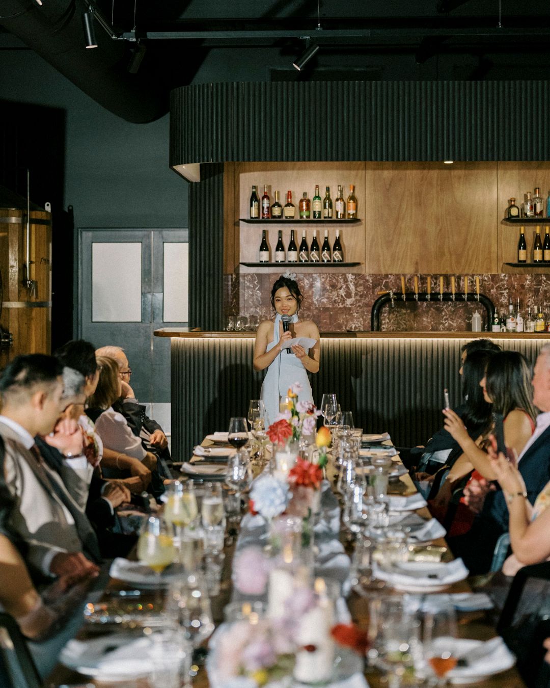 Woman speaking at microphone during elegant restaurant dinner event with guests seated at long decorated table.