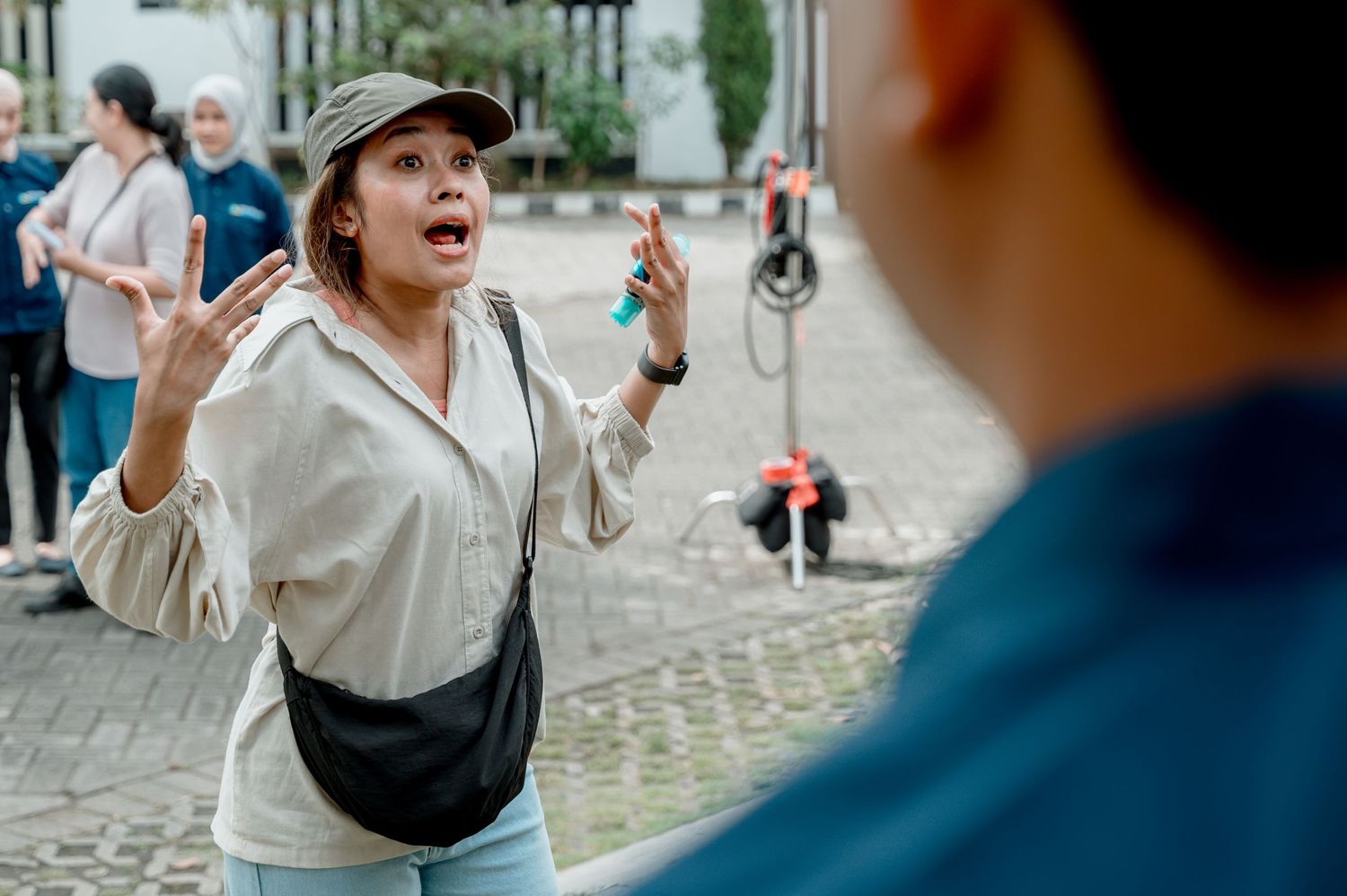A woman in a cap and crossbody bag gestures expressively while speaking outdoors on a cobblestone plaza.
