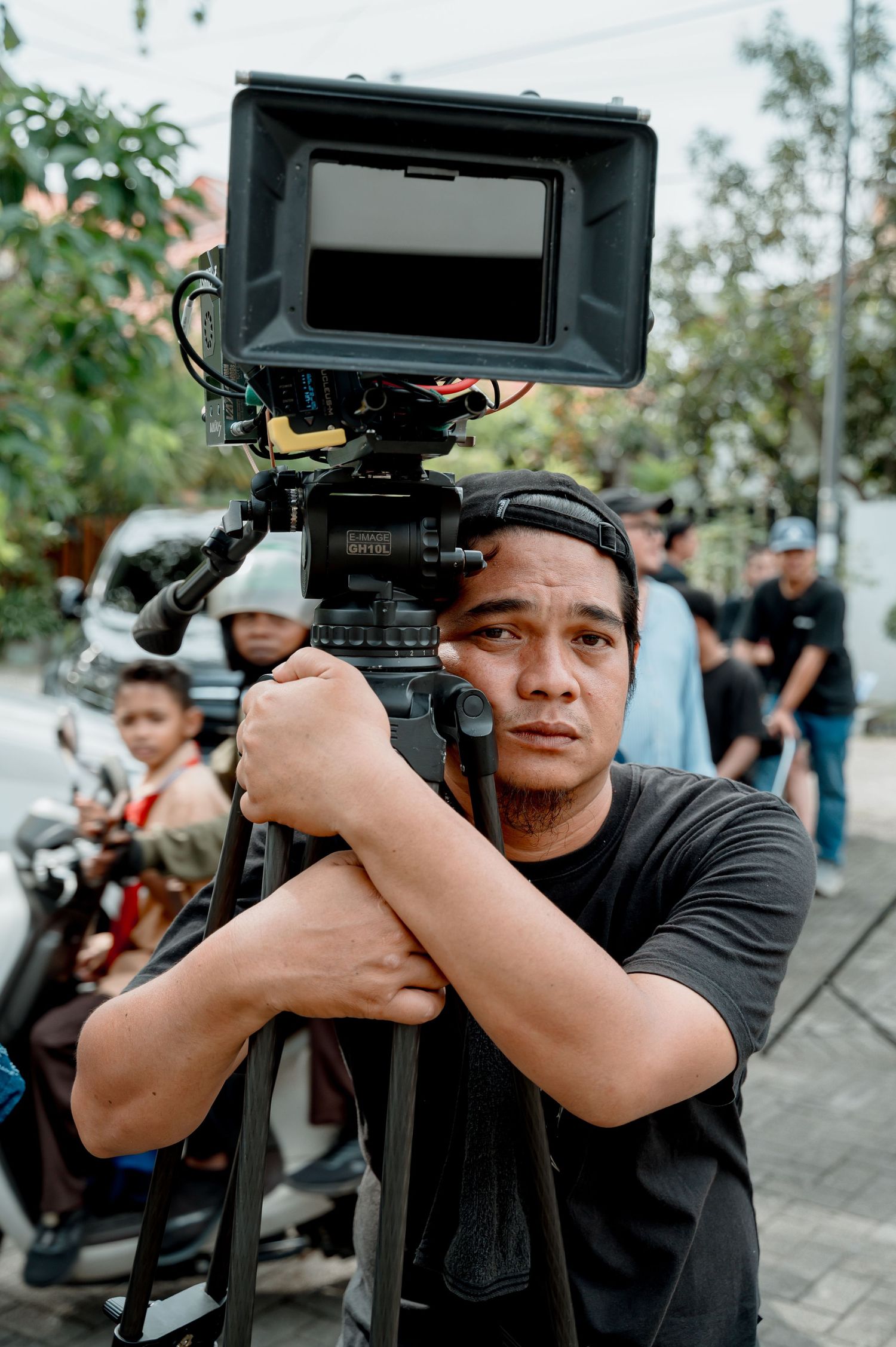 A cameraman in a black t-shirt operates a professional video camera on a tripod outdoors on a busy street.