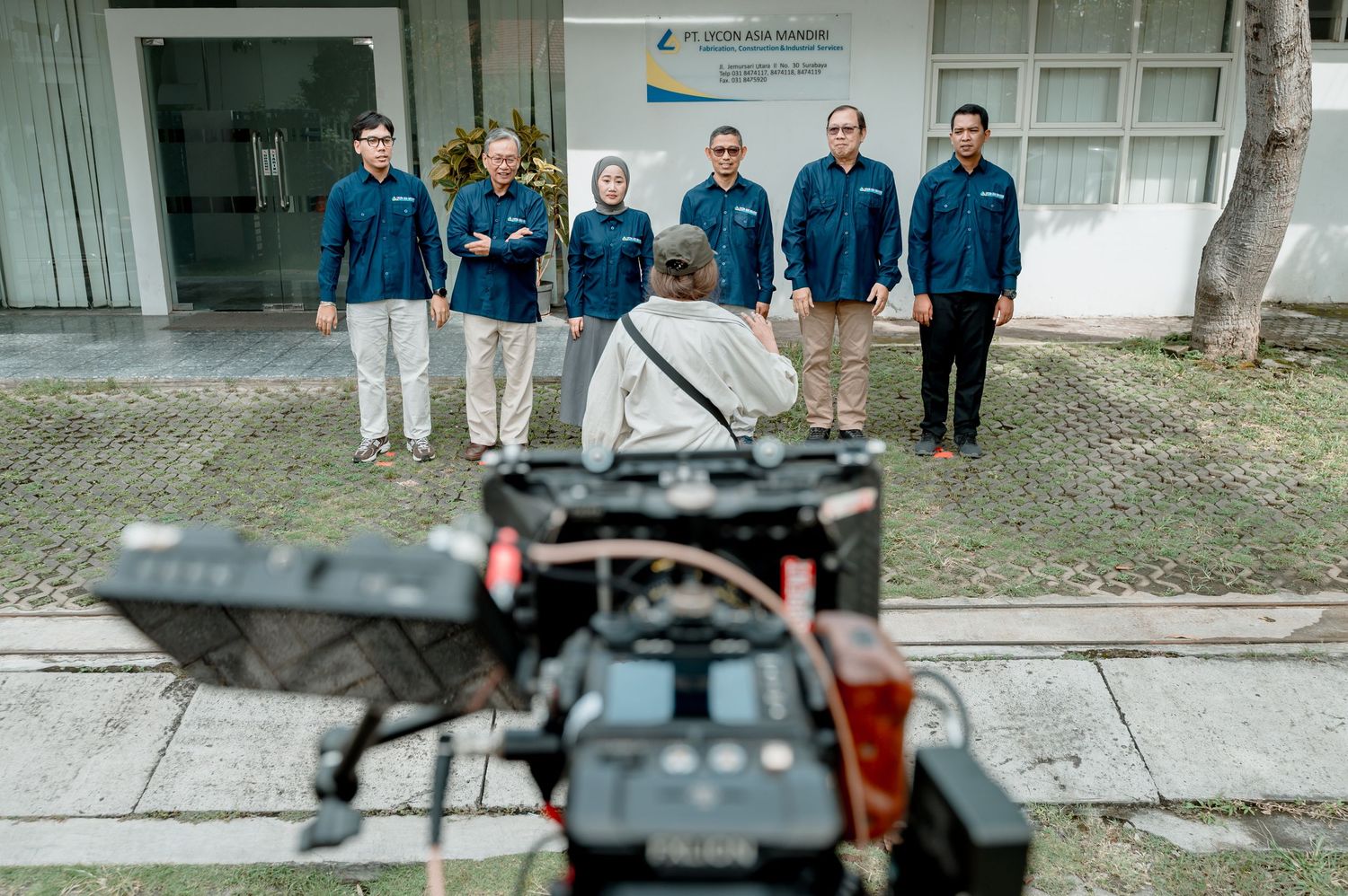 A camera crew films a group of six professionals in navy uniforms standing outside an office building.