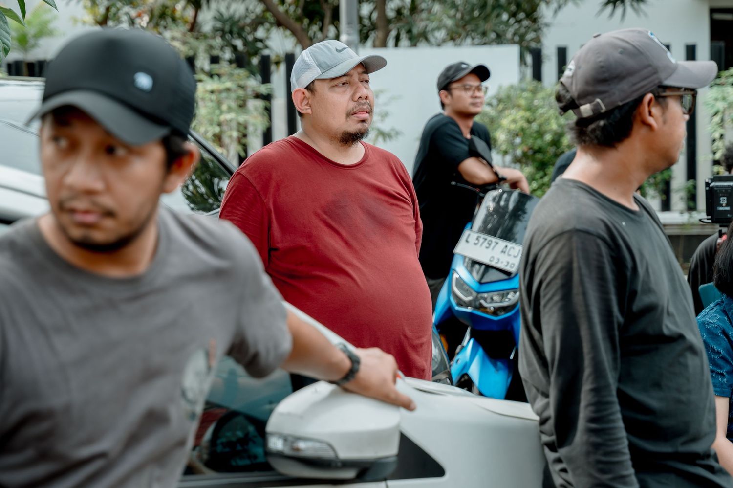 A group of men standing outdoors near vehicles, with one man in a red shirt at the center of the scene.