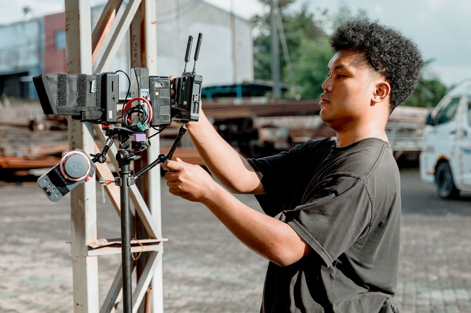 Young videographer adjusting professional camera equipment on a metal stand outdoors at a film production site.