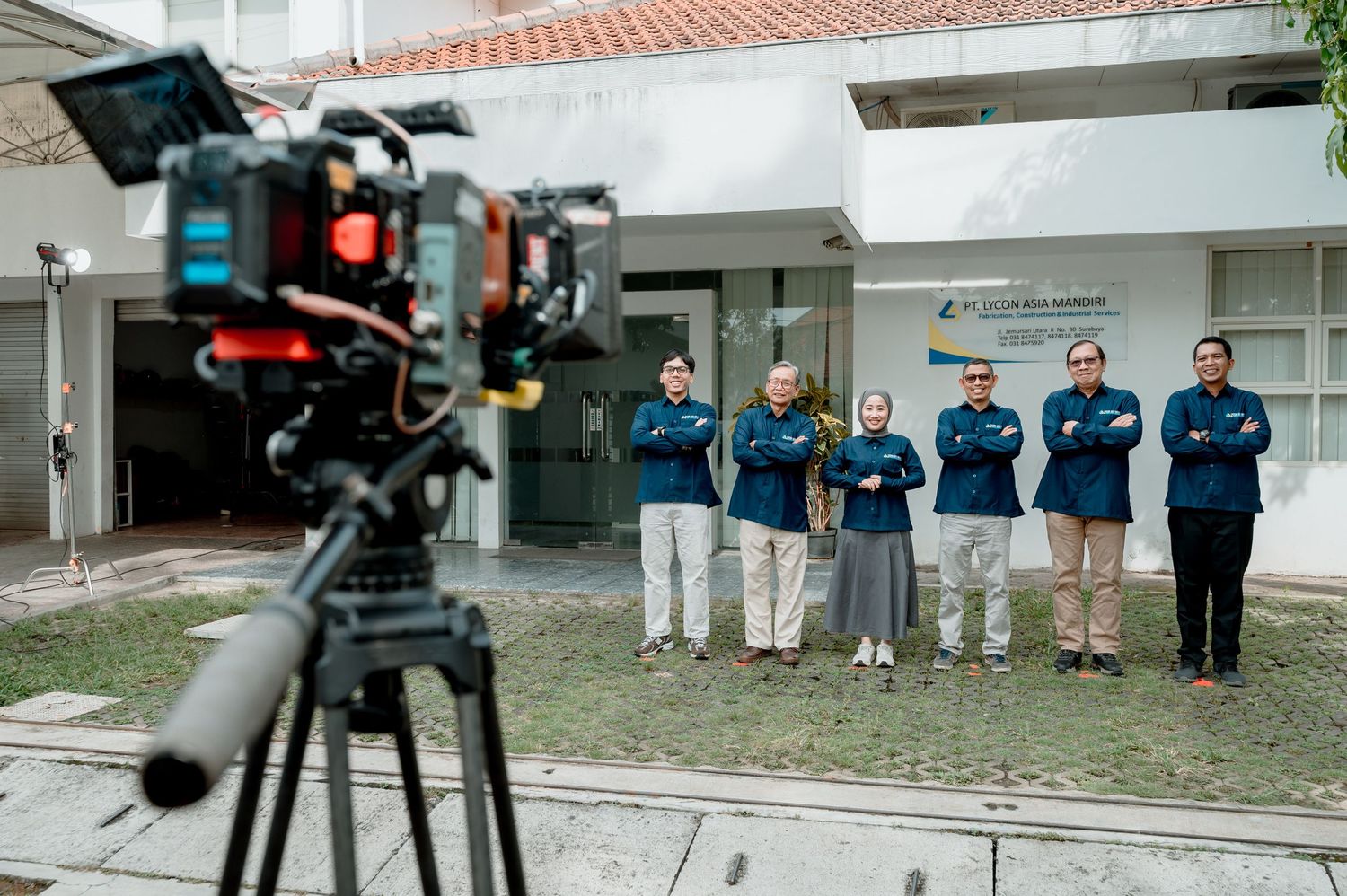 A film crew records a group of five professionals in navy jackets standing outside a modern building.