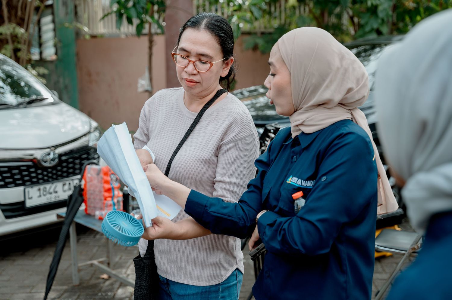 Two women reviewing documents outdoors near parked cars, one wearing hijab in blue uniform consulting with another.
