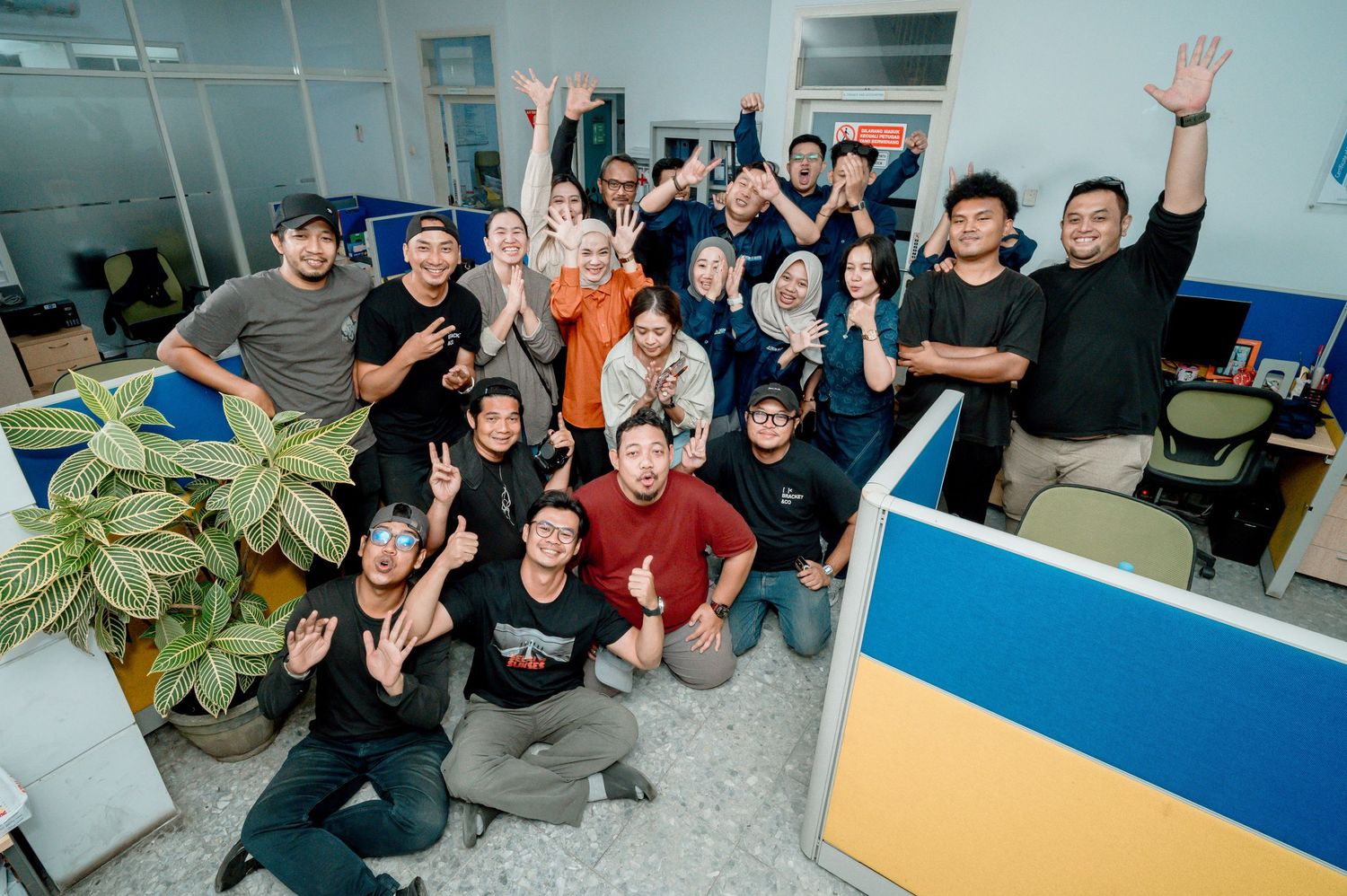 Diverse group of cheerful office workers posing and waving in a modern workspace, celebrating team success together.