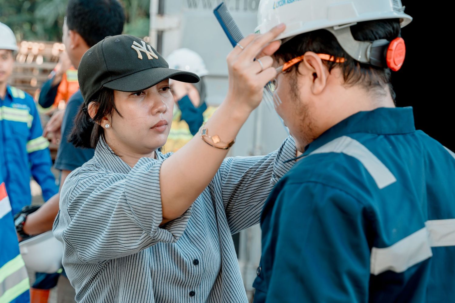 Woman in NY cap adjusting hard hat on male worker in blue uniform at industrial worksite.