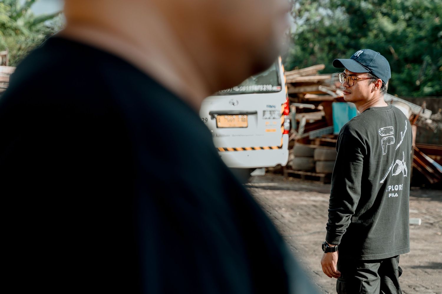 Two men in black long-sleeve shirts stand in an outdoor scrapyard, one wearing a blue cap.