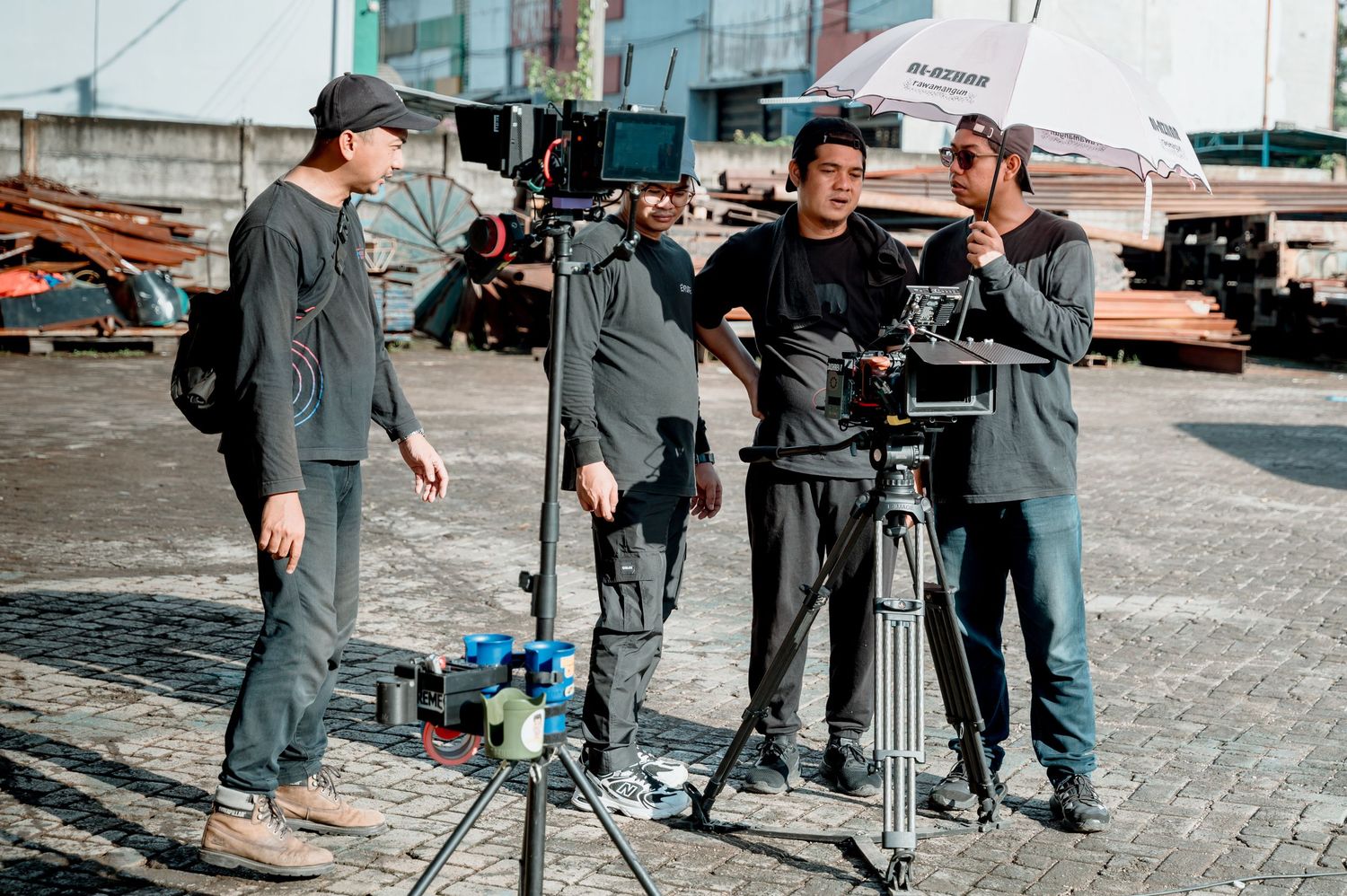 Film crew with camera on tripod shooting outdoors, one holding umbrella, in an industrial setting.