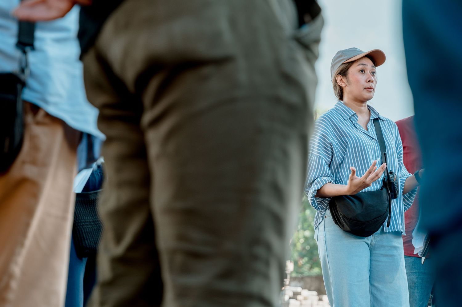 A woman in a striped shirt and cap gestures while speaking to a group outdoors.