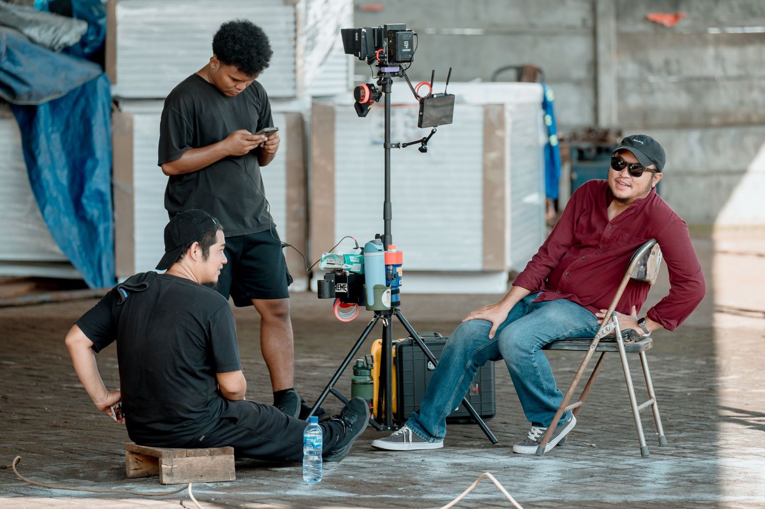 Three film crew members on set, two in black shirts and one in red relaxing on a folding chair near camera equipment.