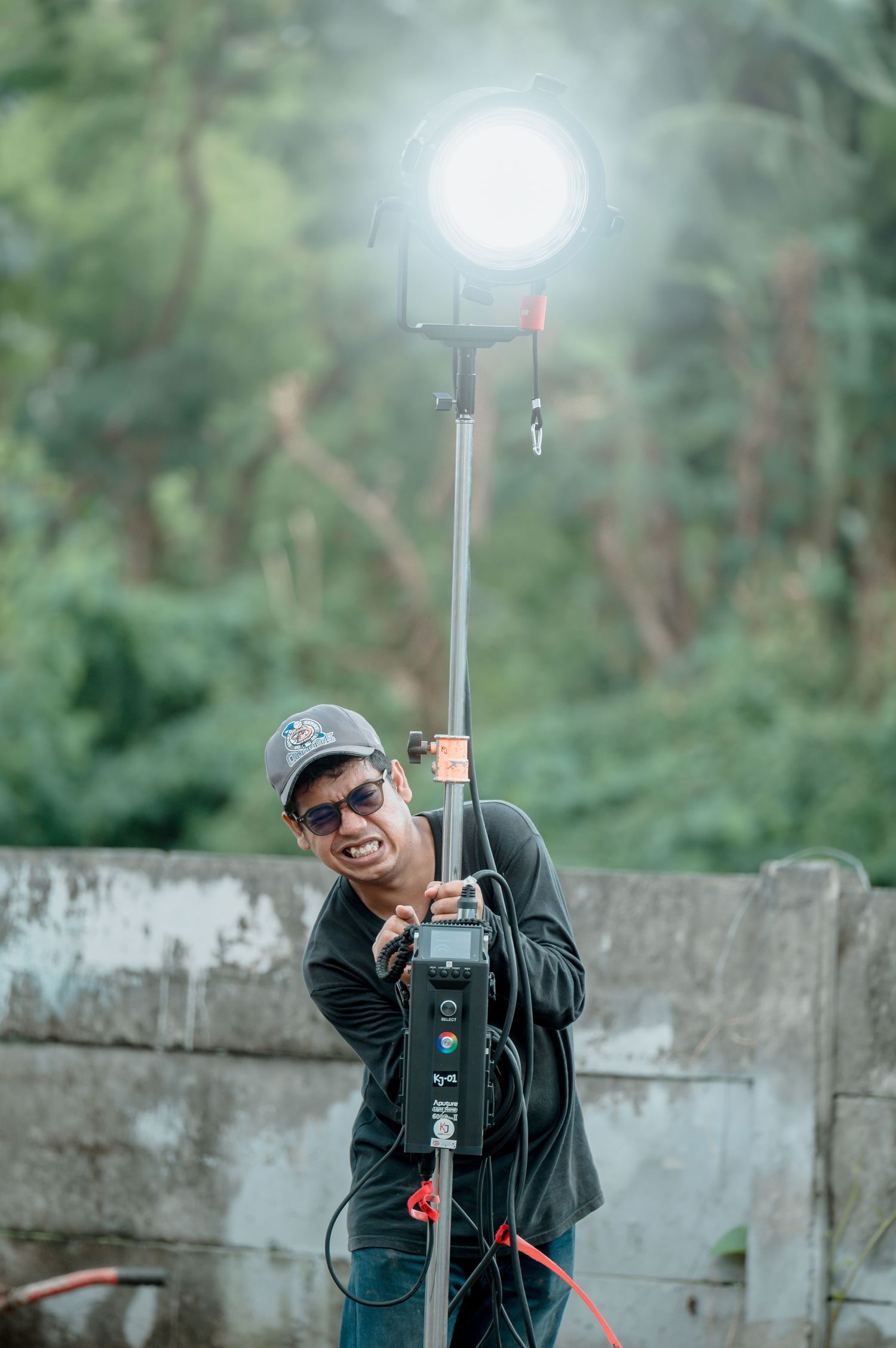 A smiling man in sunglasses and cap holds a bright studio light on a pole outdoors with green trees in background.