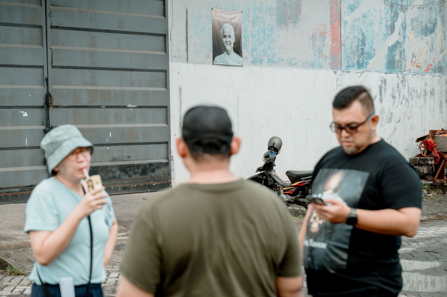 Three people standing outside near a wall with a framed portrait, one holding a drink and another checking a phone.