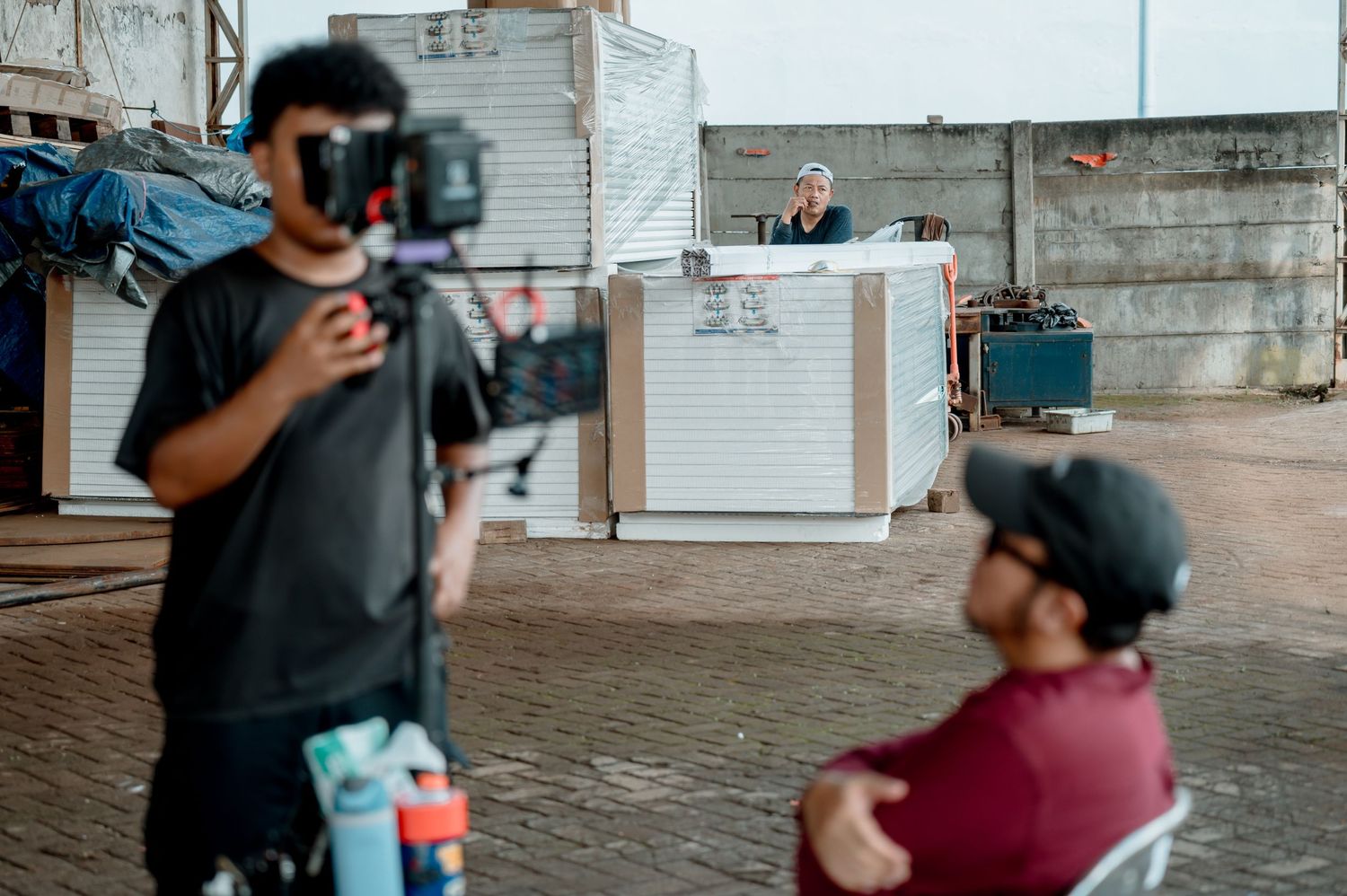 Cameraman filming an outdoor interview scene with a person seated at a desk near a shuttered building.