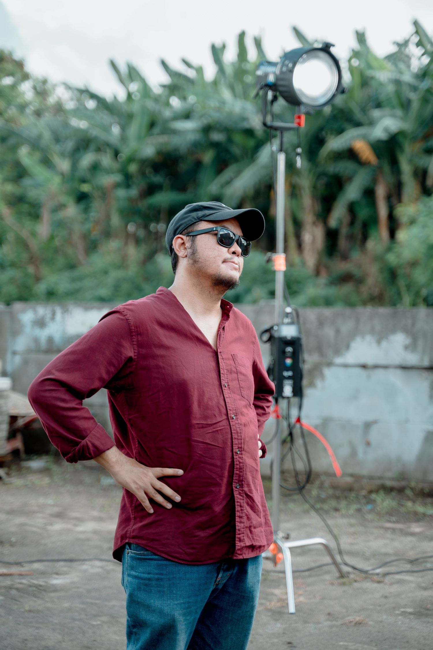 Man in red shirt, cap, and sunglasses poses outdoors near film lighting equipment with tropical trees in background.