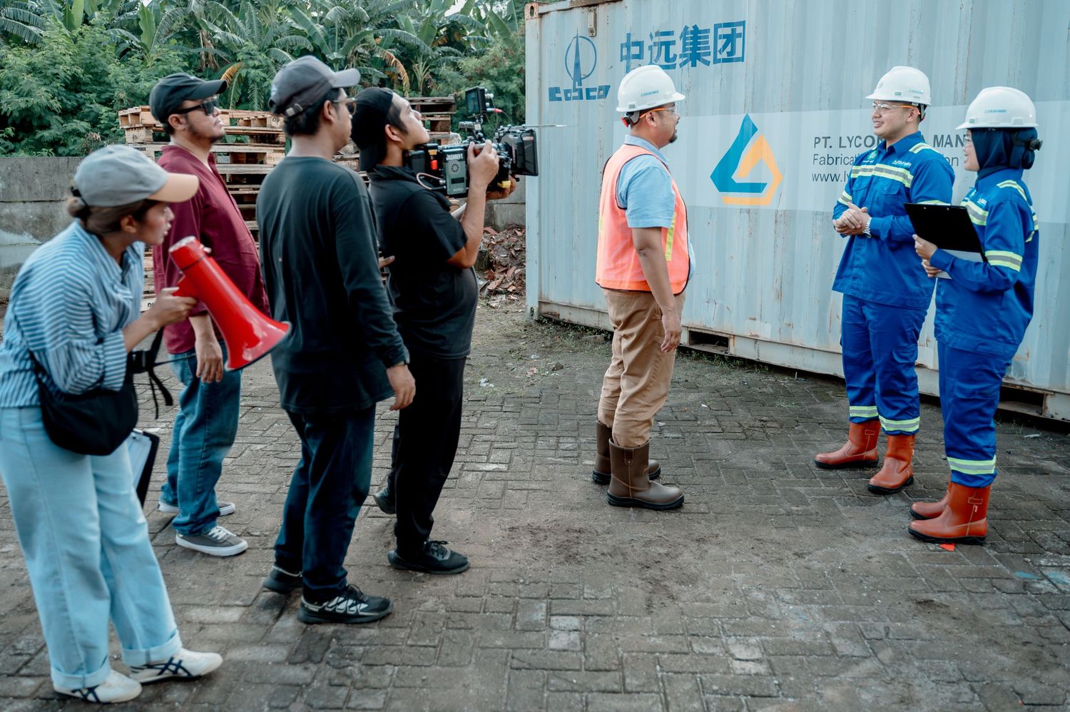 Film crew with camera and megaphone interviewing workers in hard hats and safety gear near industrial containers.