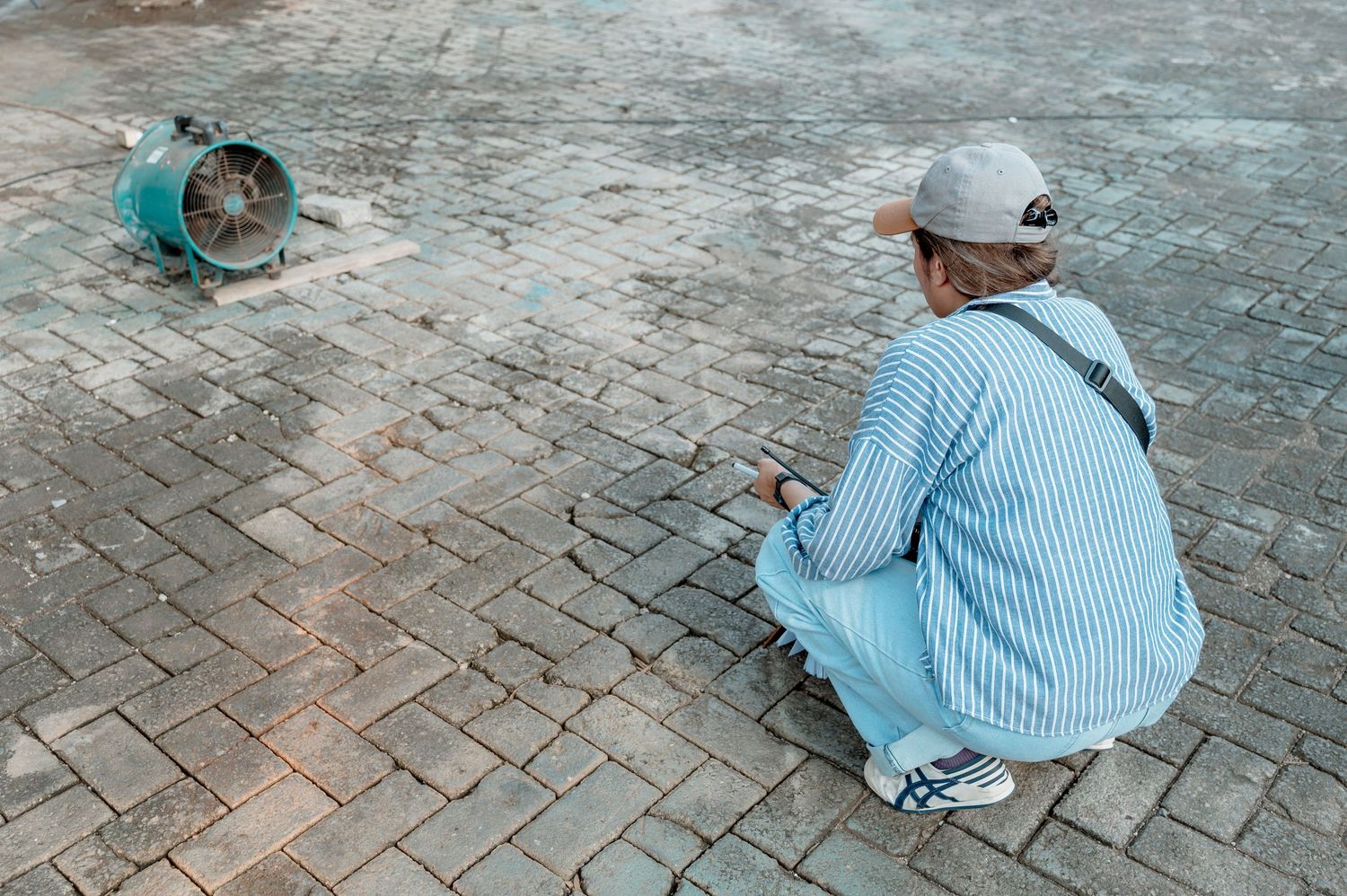 Person in striped shirt and cap crouching on brick pavement next to a green industrial fan outdoors.